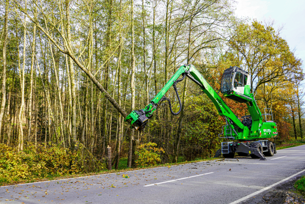 SENNEBOGEN 718 E Fällbagger mit Greifersäge bei Baumpflegearbeiten an einer bewaldeten Straße.