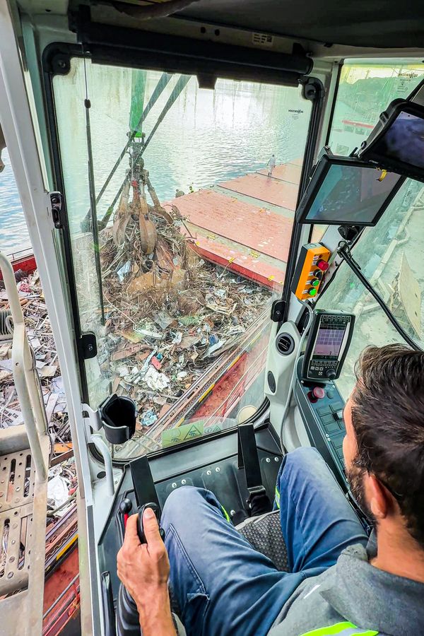 Blick aus der SENNEBOGEN Maxcab-Fahrerkabine auf den Mehrschalengreifer beim Entladen von Metallschrott aus einem Schiff.